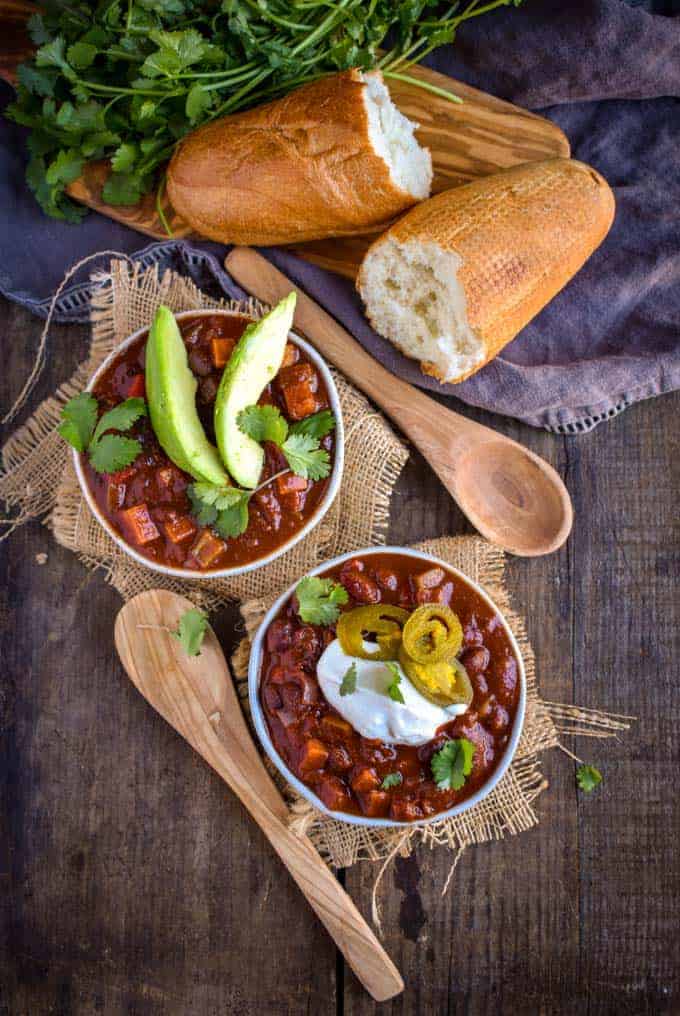 Two bowls of sweet potato black bean chili on table with bread and spoons.