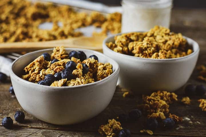Two bowls of healthy oil-free granola on wooden table with blueberries and milk.