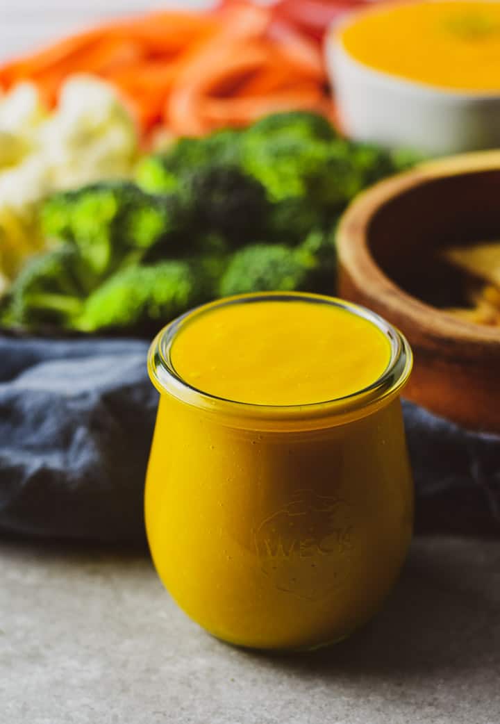 Vegan cheese sauce in jar on table in front of veggies.