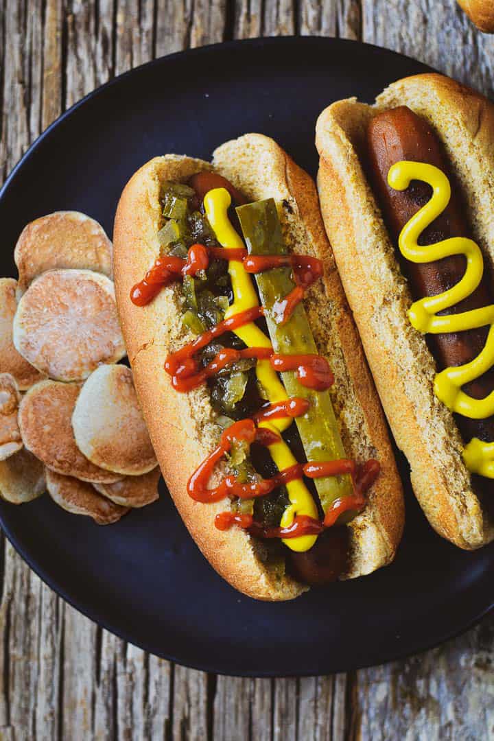 Vegan carrot dog with relish, ketchup, mustard, and pickle and one with mustard only on plate with chips.