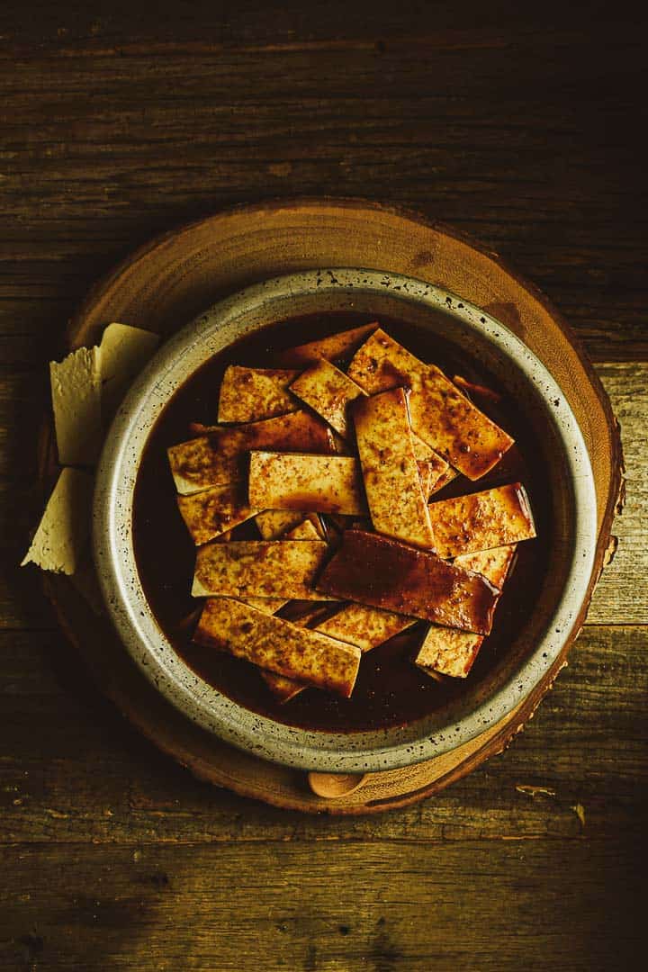 Sliced tofu in marinade in metal bowl on cutting board.