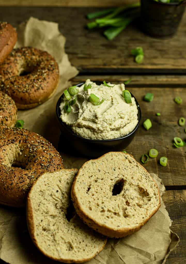 Vegan cream cheese in black bowl with chopped green onions and surrounded by bagels.