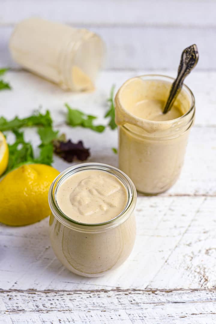 Two jars of garlicky lemon tahini dressing on a table with lemon and salad leaves.