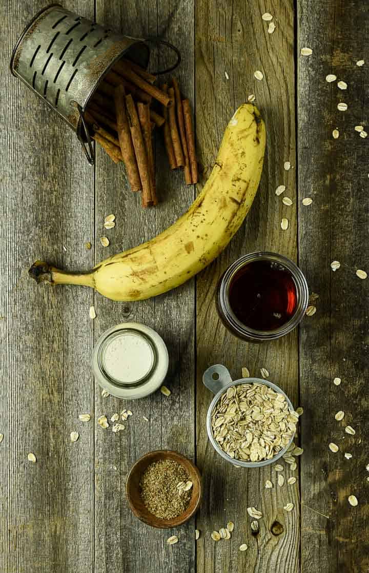 Banana, oats, cinnamon stick, maple syrup, milk, and flax on table.