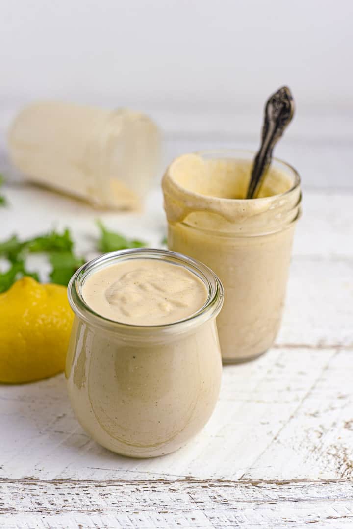 Lemon tahini dressing in two jars on white table with lemons and greens.