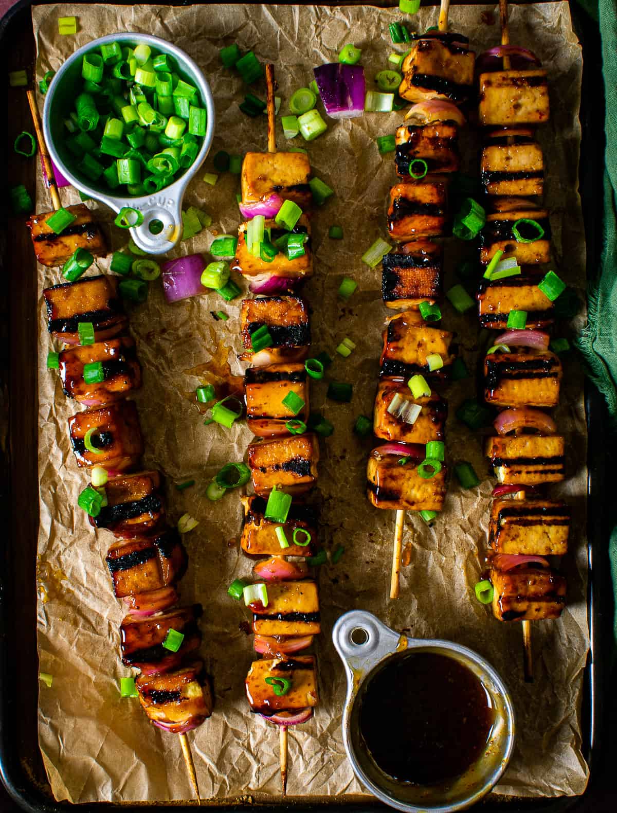 grilled teriyaki tofu skewers on parchment paper with chopped green onion and teriyaki sauce.